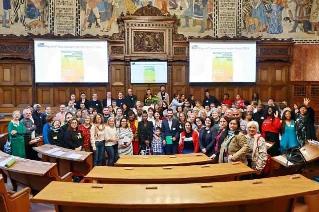 Ein Gruppenbild im Basler Grossen Rat. Die Teilnehmenden der Migrant*innensession stehen vor dem Rednerpult, vor ihnen sind ein paar Sitzbänke im Bild. 