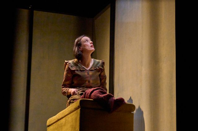 Eine Frau sitzt auf einem schmalen Bett in einem spärlich beleuchteten, kargen Raum mit hohen, beigefarbenen Wänden. Sie trägt ein schlichtes, historisch wirkendes Kleid und blickt mit angespanntem, nachdenklichem Ausdruck nach oben. Das Bühnenlicht hebt ihre Figur hervor und erzeugt eine ruhige, zugleich bedrückende Atmosphäre.