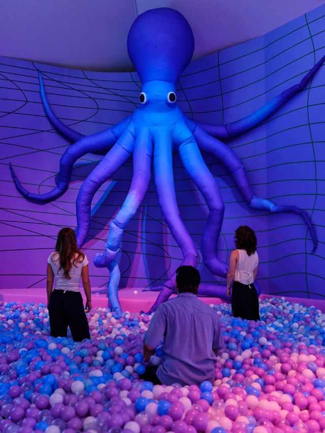 Ausstellungsansicht, drei Menschen sind im Bällebad und schauen einen grossen Oktopus an. Die Farben sind Blau, Lila und Pink.