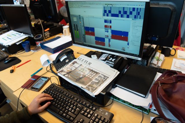 Ein Schreibtisch in einem Büro mit einem Computerbildschirm, auf dem ein Audiobearbeitungsprogramm mit mehreren Tonspuren zu sehen ist. Vor dem Monitor liegen eine Zeitung mit der Überschrift „Kultur“, ein Telefon, ein Smartphone, Stifte und verschiedene Papiere. Eine Person tippt auf einer Tastatur, während auf dem Tisch außerdem ein Notizbuch und eine Handtasche liegen.