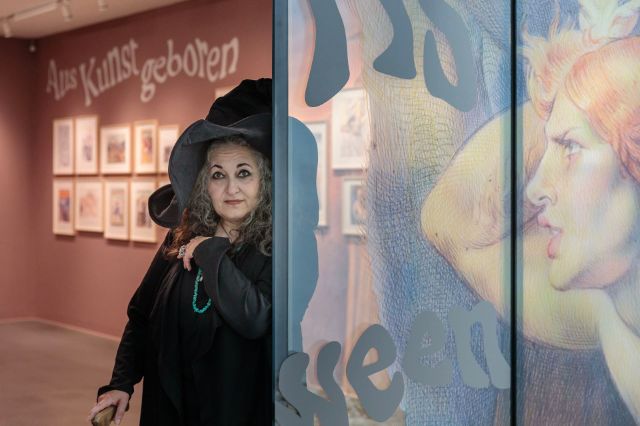 Emil Ferris trägt einen Hexenhut und schwarze Kleidung, sie hält einen Gehstock. Sie steht am Anfang der Ausstellung, im Hintergrund sind Bilder an der Wand zu sehen.