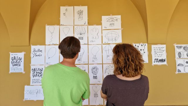 Zeichnungen von Ballons hängen an der Wand. Marcel Hörler und Michèle Degen stehen davor und schauen die Wand an.