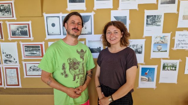Marcel Hörler steht links und Michèle Degen rechts vor einer gelben Wand, darauf sind Archivbilder zu sehen.