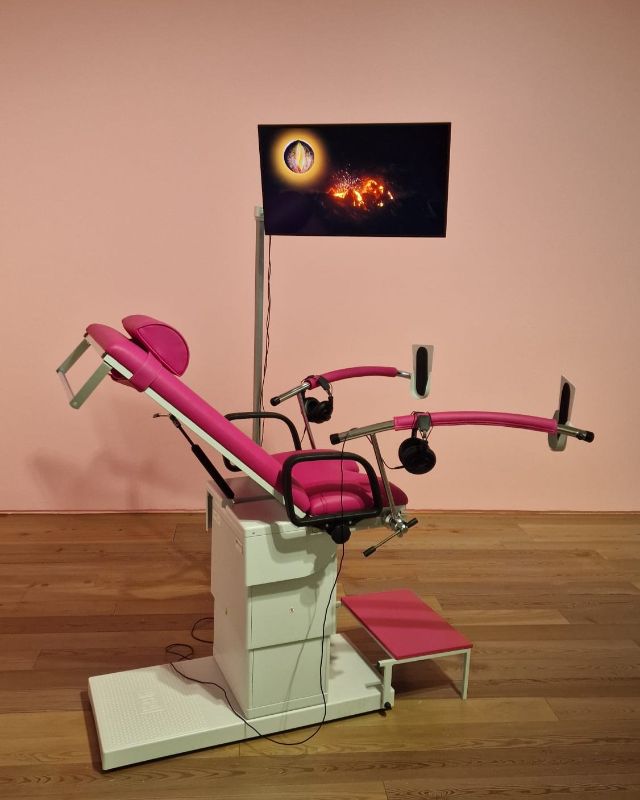 Ein pinker Gynäkologie-Stuhl, daran befestigt ist ein Bildschirm, auf dem ein Video läuft.