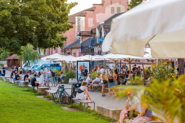 Der Eingang der Kaserne mit Menschen im Sommer unter Sonnenschirmen