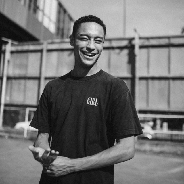Loye Carner auf einem Schwarz-Weissbild vor einer Industriehalle am Lächeln