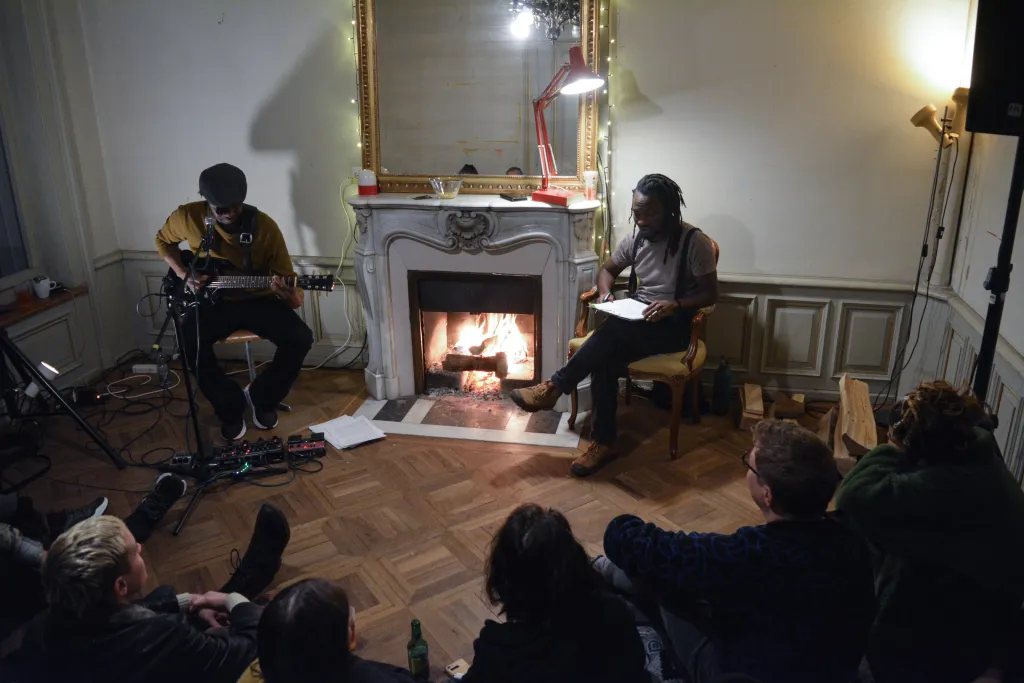 Zwei Menschen sitzen links und rechts neben einem Cheminée, spielen Gitarre und präsentieren einen Text. Zuschauende sitzen im Halbkreis vor ihnen und hören gespannt zu.