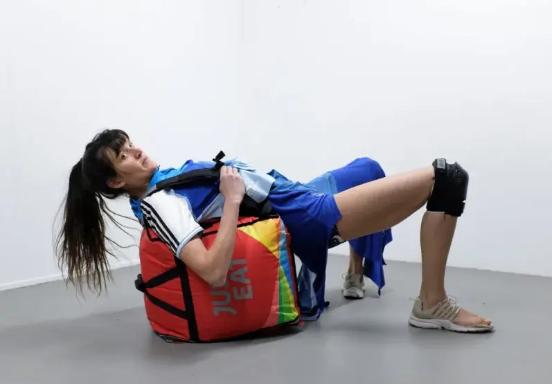Eine Frau in Sportkleidung lehnt sich auf einen Just Eat Delivery Rucksack. Die Füsse sind auf dem Boden platziert, die Hüfte in der Luft, der Rücken gestützt vom Rucksack auf dem Boden. Sie schaut zur Kamera.